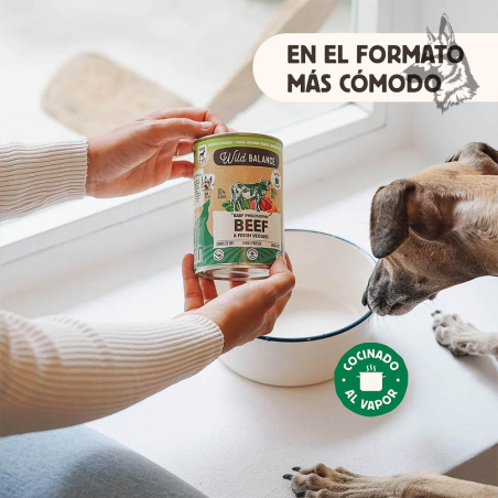 Una persona sostiene una lata de Wild Balance Beef & Fresh Veggies mientras un perro la observa junto a su comedero, resaltando 